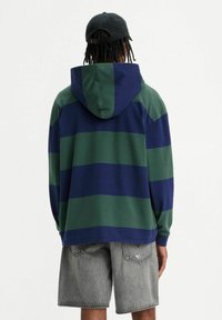 Persona con capelli intrecciati che indossa un berretto nero al contrario, una felpa a righe blu e verde e pantaloni corti di jeans grigi, girata di spalle su uno sfondo neutro.