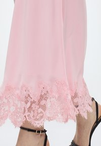 Πόδια με μαύρα σανδάλια με λουράκι στον αστράγαλο, εμφανή κάτω από μια ροζ φούστα με λεπτομερή δαντέλα με σχέδιο λουλουδιών στο τελείωμα, σε λευκό φόντο.