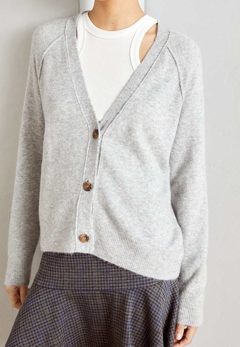 Femme portant un cardigan gris clair boutonné sur un débardeur blanc et une jupe à carreaux aux tons atténués.