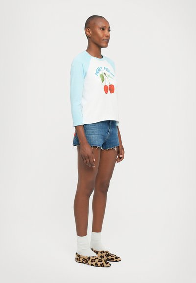 Woman wearing light blue-sleeved shirt with cherry graphic, denim shorts, white socks, and leopard print flats, standing on white background.