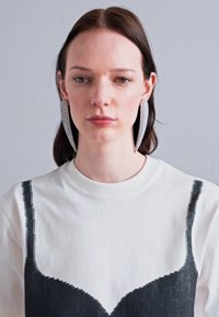 Dlhé strieborné náušnice s textúrovaným, predĺženým dizajnom. Model má na sebe biele mikinu vrstvenú cez čierny top.