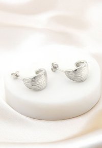 Silverfärgade hängörhängen med en texturerad yta, i en böjd form med post-back stängning, visade på en slät vit pelare.