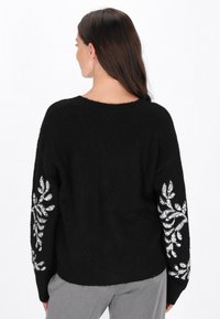 Fekete pulóver hosszú, laza szabással. Ezüst levél mintákkal díszített mindkét ujján. Texturált, puha anyagból készült.
