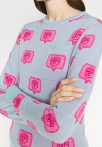 Ljusblå stickad tröja med rosa ögonformade mönster och en liten bröstficka. Mudden och halsringningen är i en kontrasterande ljusblå färg.