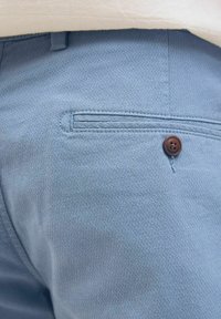 Šviesiai mėlynos tekstūruotos kelnės su galine kišene, su ruduo mygtuko akcentu ir siuvinėtų detalių. Audinys rodo subtilų pynimo raštą.