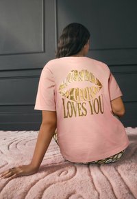 Šviesiai rožinė medvilninė marškinėlių palaidinė su auksiniu metaliniu užrašu „ROCKETT ST. GEORGE MYLI TAVE“ ir lūpų atspaudu ant nugaros.