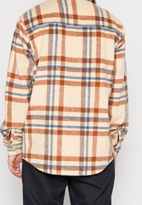 Osoba w kremowej, rdzawoczerwonej i niebieskiej koszuli w kratę z długim rękawem i zapiętymi mankietami, pokazana od tyłu, w połączeniu z czarnymi spodniami.