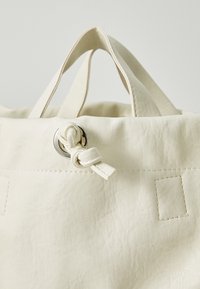 Hvit tote-veske med teksturert finish, knuteavslutning med metallbeslag og faste håndtak for bæring.