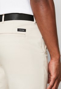 Pantaloni in cotone beige con una cintura in tessuto nera. Presentano una tasca posteriore con una piccola etichetta nera con la scritta "Calvin Klein." Texture liscia.