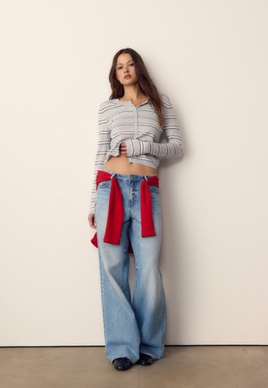 Mladi žena nosi prugasti gornji dio s gumbima, široke svijetloplava traperice i crveni džemper vezan oko struka, stoji uz običan zid.