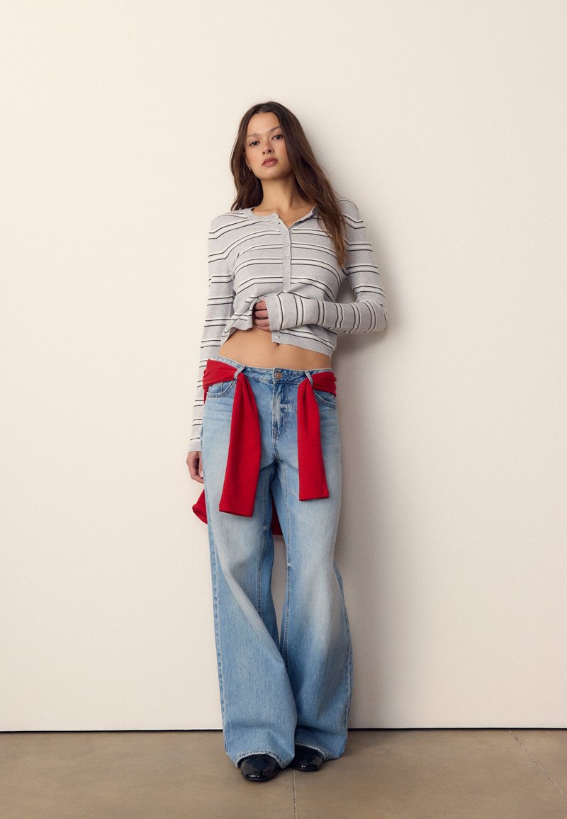 Jeune femme portant un crop top rayé boutonné, un jean large bleu clair et un pull rouge noué autour de la taille, debout devant un mur uni.