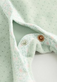 Lys grøn stof heldragt med små polkaprikker, blomsterprint detaljer og sølv- samt kobberknapper til lukning. Blød tekstur, hyggeligt materiale.