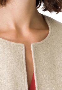 Beige gemêleerd stoffen jack met een ronde halslijn, met een rechte snit en een open voorkant ontwerp. Daaronder is een vleugje rood te zien.