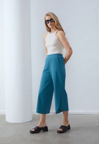 Lys beige tettsittende tank topp kombinert med teal avkuttede bukser og svarte sklitesandaler. Bukser har en strukturert, flytende design.