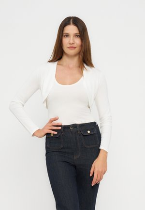 Femme aux cheveux bruns raides portant un cardigan court blanc, un débardeur blanc et un jean taille haute bleu foncé, debout avec une main sur la hanche.