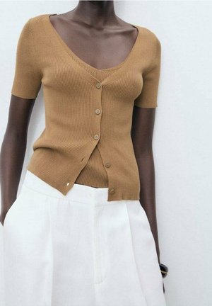Personne à la peau foncée portant un haut côtelé marron clair à manches courtes boutonné et un pantalon blanc taille haute avec les mains dans les poches.