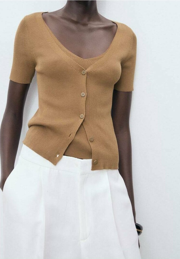 Personne à la peau foncée portant un haut côtelé marron clair à manches courtes boutonné et un pantalon blanc taille haute avec les mains dans les poches.