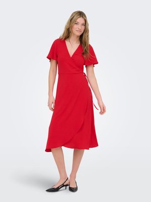 Tânără stând în picioare, purtând o rochie roșie tip wrap cu mâneci evazate și pantofi negri slingback cu toc mic, pe fundal alb.