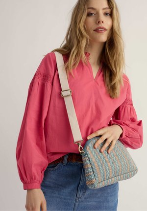 Donna con capelli biondi sciolti che indossa una blusa rosa a maniche lunghe e jeans, che tiene una borsa a tracolla a righe blu e pesca con texture.