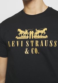 Camiseta de algodón negra con un diseño gráfico dorado que muestra caballos y figuras, con la leyenda "LEVI STRAUSS & CO." y "PATENTADO MAYO 1873."