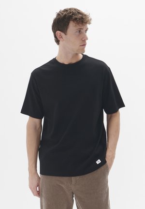 T-shirt noir à manches courtes en coton, avec col rond et une petite étiquette blanche sur le côté inférieur gauche. Texture lisse, coupe décontractée.