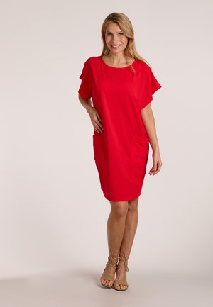 Woman wearing a loose red knee-length dress with pockets, smiling, standing with one hand on hip and beige strappy sandals.