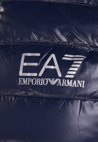 Giacca puffer blu navy con finitura lucida, con un logo grigio prominente: "EA7 Emporio Armani" sulla parte frontale.