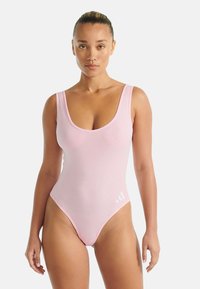 Kvinna med bakåtkammat hår som bär en ljusrosa ärmlös bodysuit med en liten vit logotyp på vänster höft, stående mot en enfärgad bakgrund.