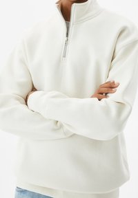 Felpa zip con colletto alto di colore crema chiaro, realizzata in un morbido tessuto texturizzato, con zip a un quarto e polsini a costine. Vestibilità casual.