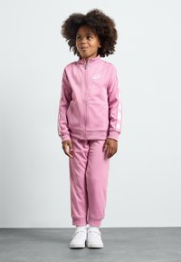 Rosa treningsdrakt laget av mykt stoff, med en zip-up jakke med en hvit logo og matchende bukser med mansjetter på anklene. Brukes med hvite joggesko.