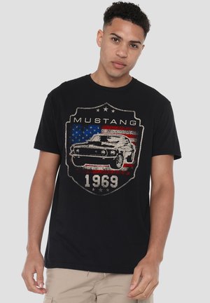 Junger Mann trägt ein schwarzes T-Shirt mit einem Vintage-Mustang-1969-Autografik über einem Hintergrund der amerikanischen Flagge und beigen Hosen.