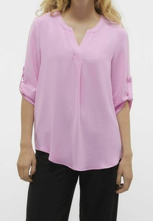 Vrouw draagt een losse lichtroze V-hals blouse met opgerolde mouwen en zwarte broek, staand voor een effen witte achtergrond.