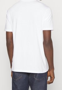 Vit bomullströja med korta ärmar, rund hals och rak fåll. Bakifrån visas en enkel, minimalistisk design.