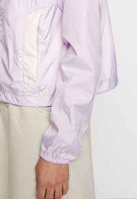 Manche de veste violet clair avec poignet élastique et détail de poche contrastant, montrée sur le bras d'une personne sur un fond uni.