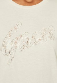 Camiseta de color beige claro con un logo "Guess" adornado con encaje y rhinestones. Tela suave con una textura lisa y cuello redondo.
