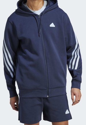 Homme portant un sweat à capuche zippé Adidas bleu marine et un short assorti avec logo blanc et bandes sur les manches, debout devant un fond uni.