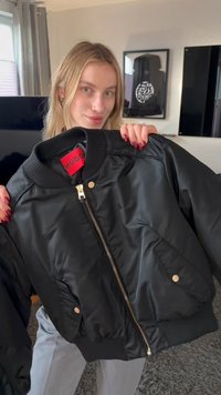 Noor naine hoiab käes musta Hugo pommitaja jopet kuldsete tõmblukkude ja nuppudega, seistes moodsas elutoas, kus on raamitud seinamaalid.