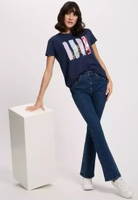 Granatowa koszulka z krótkim rękawem z kolorowym wzorem graficznym. W zestawie z dżinsami w wysokim stanie w kształcie dzwonów oraz białymi trampkami.
