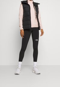 Černá vatovaná vesta přes světle růžový fleecový zip, doplněné černými legínami s bílým logem a bílými teniskami s texturovanými podrážkami.