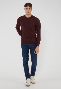 Maglione bordeaux a maniche lunghe con collo rotondo, realizzato in tessuto morbido. Jeans blue slim fit e sneakers bianche con suola beige. Logo discreto sulla manica.