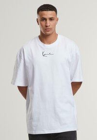 White cotton T-shirt with short sleeves and a relaxed fit. Features a black signature logo on the left chest. Smooth texture, simple design.