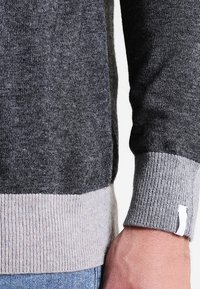 Grå stickad tröja med en mörkgrå kropp, ljusgrå ribbade muddar och en liten vit etikett vid fållen. Mjukt material och figurpassande design.