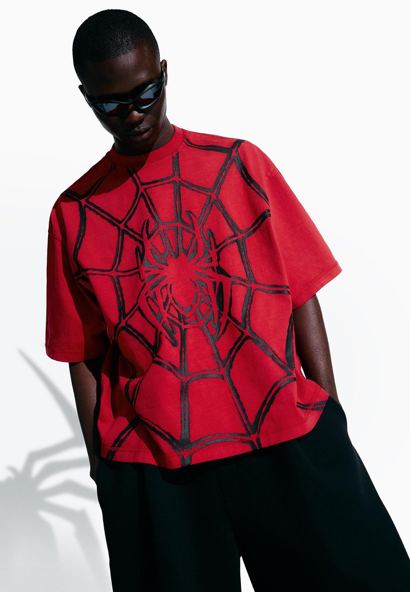 Junger Mann mit schwarzen Sonnenbrillen, rotem, übergroßem T-Shirt mit schwarzem Spinnen- und Netzdesign und schwarzen Hosen, dessen Schatten die Form einer Spinne annimmt.