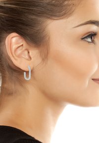 Boucles d'oreilles en métal argenté avec un design en boucle ouverte, présentant une texture lisse et une forme minimaliste, portées sur l'oreille d'un mannequin.