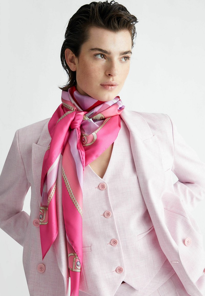 Pañuelo de seda rosa con una textura suave, que presenta un degradado de tonos rosas y un patrón de cadenas, atado alrededor de un cuello debajo de un traje rosa claro.