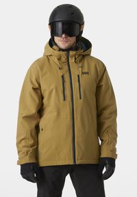 Giacca da sci marrone con cappuccio, cerniere nere e due tasche frontali. Finitura opaca, indossata con casco nero e guanti.