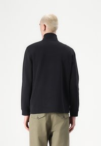 Schwarzer Langarmpullover mit hohem Kragen und gerippten Bündchen, aus einem glatten Stoff gefertigt, kombiniert mit hellgrünen Hosen mit Gesäßtaschen.