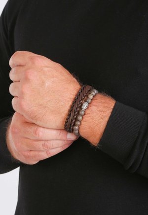 Vlechtarmband van bruin leer met zilveren accent en grijze kralen, gedragen om een pols, waarbij de textuur en het materiaalcontrast worden getoond.