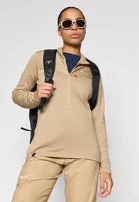 Μπεζ πλεκτό πουλόβερ με μακριά μανίκια και φερμουάρ μέχρι το ένα τέταρτο, υφασμάτινη υφή και πορτοκαλί λεπτομέρεια. Συνδυάζεται με παντελόνι της ίδιας απόχρωσης και μαύρο σακίδιο.