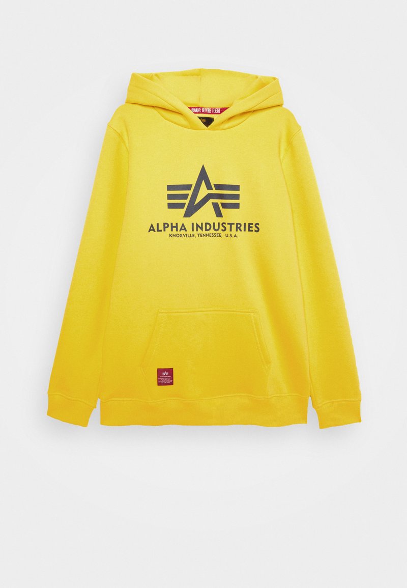 Sudadera amarilla con capucha y bolsillo tipo canguro en el frente. Presenta un logo negro "ALPHA INDUSTRIES" y una etiqueta en la parte inferior izquierda.